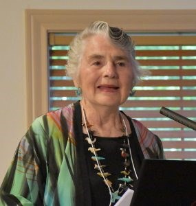 Nancy Bartlit White woman with short wavy salt and pepper hair, wearing a multicolored shawl and a necklace with many small stone birds.