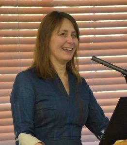 White woman with shoulder length light brown hair wearing a blue shirt standing at a microphone.