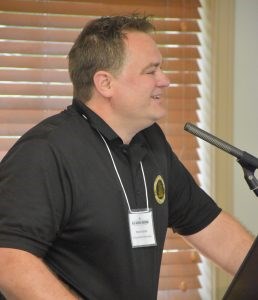 A white man with short brown hair wearing a black polo shirt standing at a microphone.