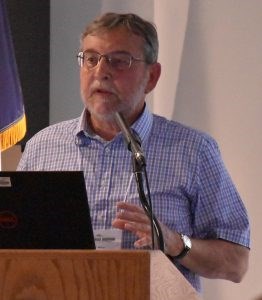 White male with gray hair, short beard, and mustache wearing pale blue plaid shirt standing at a podium.