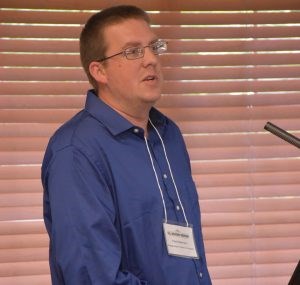 White male with glassed and short brown hair wearing a dark blue button up shirt. A nametag hangs from his neck.