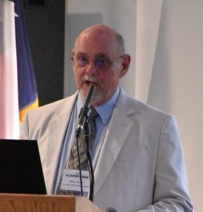 Balding white male with glasses, mustache and goatee wearing a light gray jacket, blue shirt, and dark gray tie.