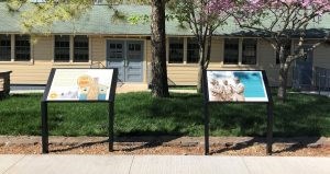 Two interpretive signs in front of tan building with nine windows and the gray doors and green shingled roof.