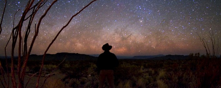 A cowboy contemplates a clear, starry night sky over the desert horizon at Big Bend National Park, Texas.