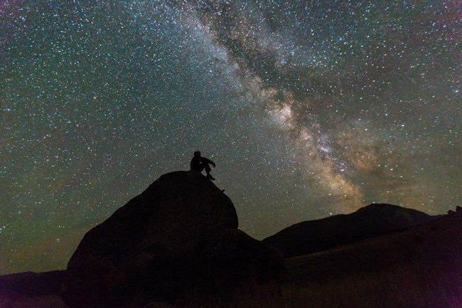 A man shown in silhouette against a brilliant night sky sits in contemplation of the galaxy from his rock perch at Yellowstone National Park.