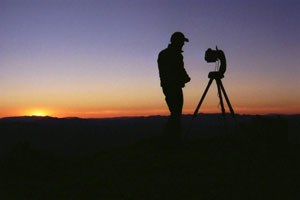 lt= An NPS scientist stands in silhouette against a dusk sky and conducts an equipment check with a CCD Camera prior to a night of data collection; Great Basin National Park. NPS / Kate Magargal.
