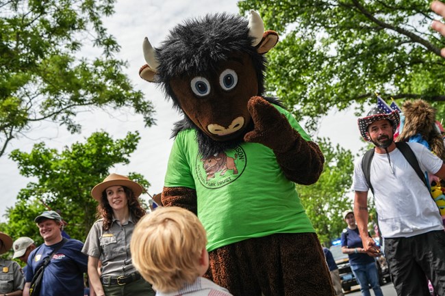 a bison mascot walks in a parade with a ranger and waves a a child