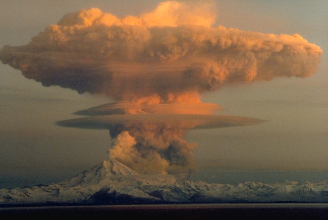 a snow capped volcano erupts with a huge ash cloud colored orange in the sunset