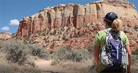 the back of a woman wearing hiking clothes and backpack in foreground with a red and tan rock cliff in the background.