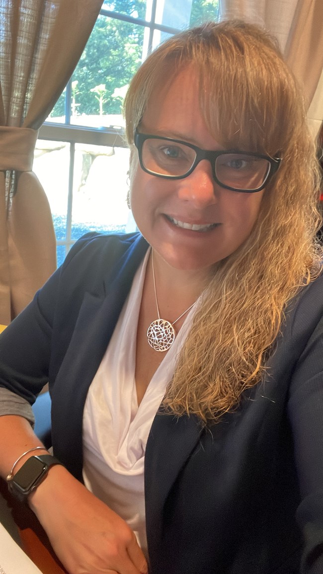 a woman with glasses and blond wavy hair smiles for the camera. She is wearing a dark colored blazer and white shirt. a woman with glasses and blond wavy hair smiles for the camera. She is wearing a dark colored blazer and white shirt.