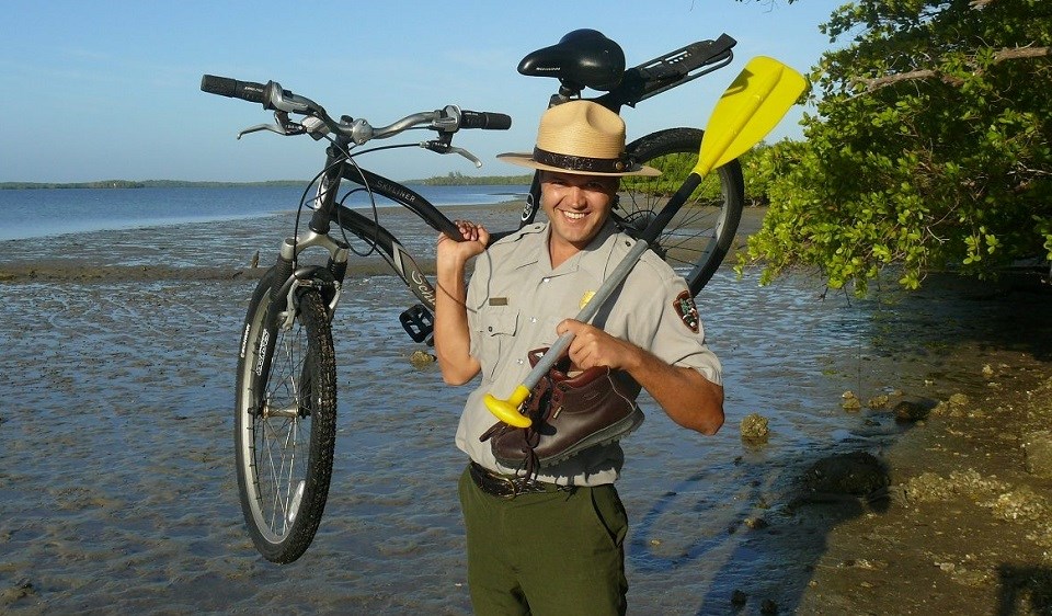 Ranger standing on a shoreline holding a bike, paddle, and hiking boat Ranger standing on a shoreline holding a bike, paddle, and hiking boat