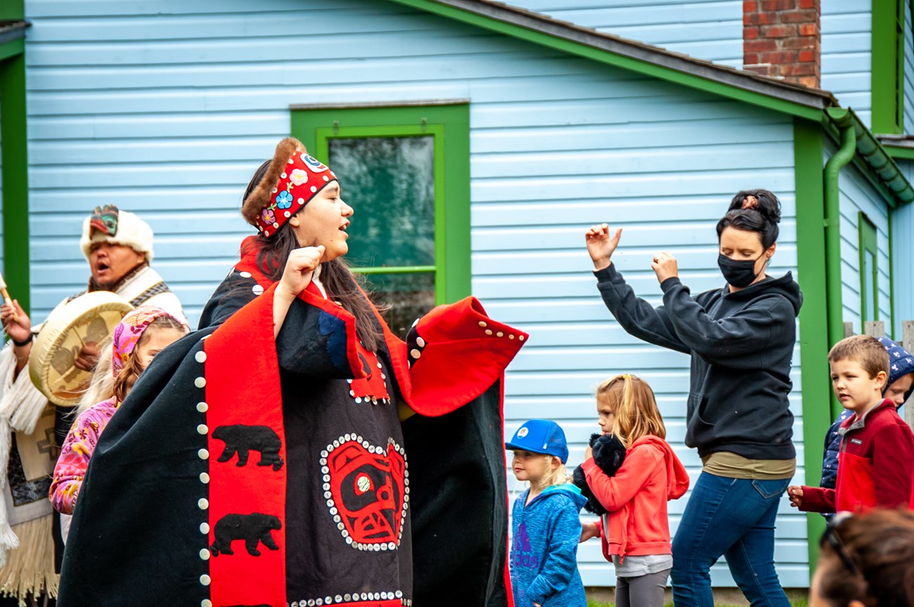 NP Gallery Junior Ranger Day KLGO Jilkaat Kwaan Heritage Dancers with a group of young children and a masked woman, dancing