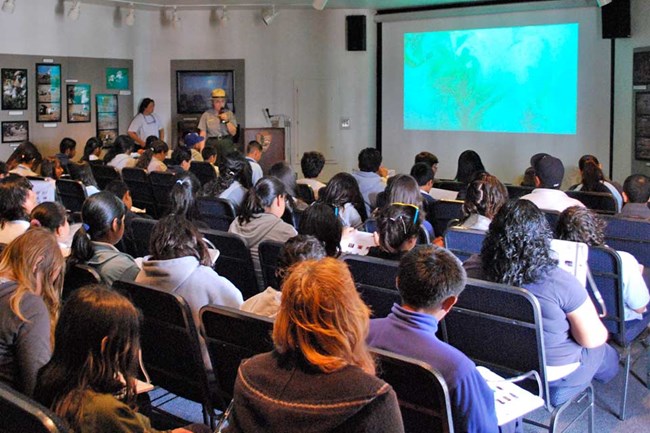 audience sitting in chairs watching video on a screen