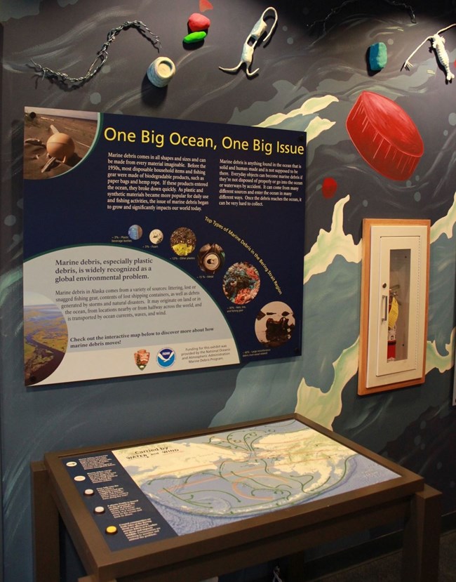 a painted mural of the ocean with 3-D trash and aquatic organisms. Within the mural there is an interpretive panel titled "One Big Ocean, One Big Issue" and below that is a map table showing the predominant currents that move marine debris in the ocean