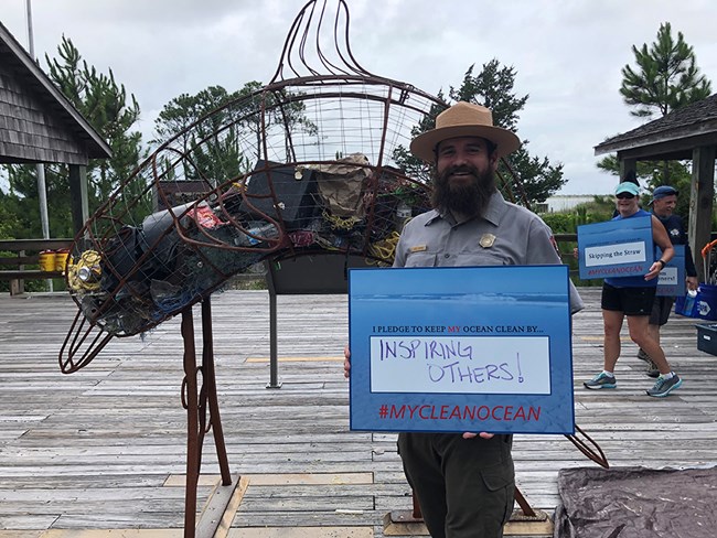 a hollow metal dolphin sculpture halfway filled with marine debris. A man in National Park Service uniform stands in front of the sculpture and holds a sign that reads "I pledge to keep my ocean clean by inspiring others!" with the hashtag #My Clean Ocean