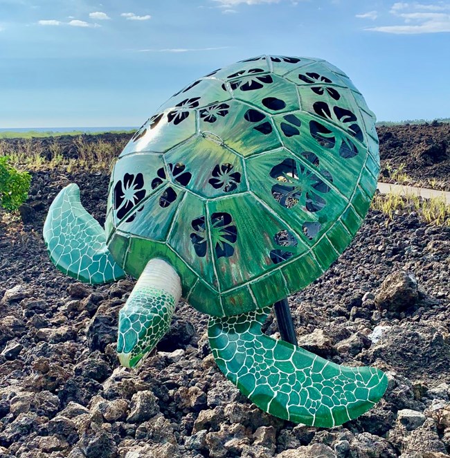 a large hollow green sea turtle sculpture with openings in the shape of flowers. The sculpture sits in a rocky lava field.