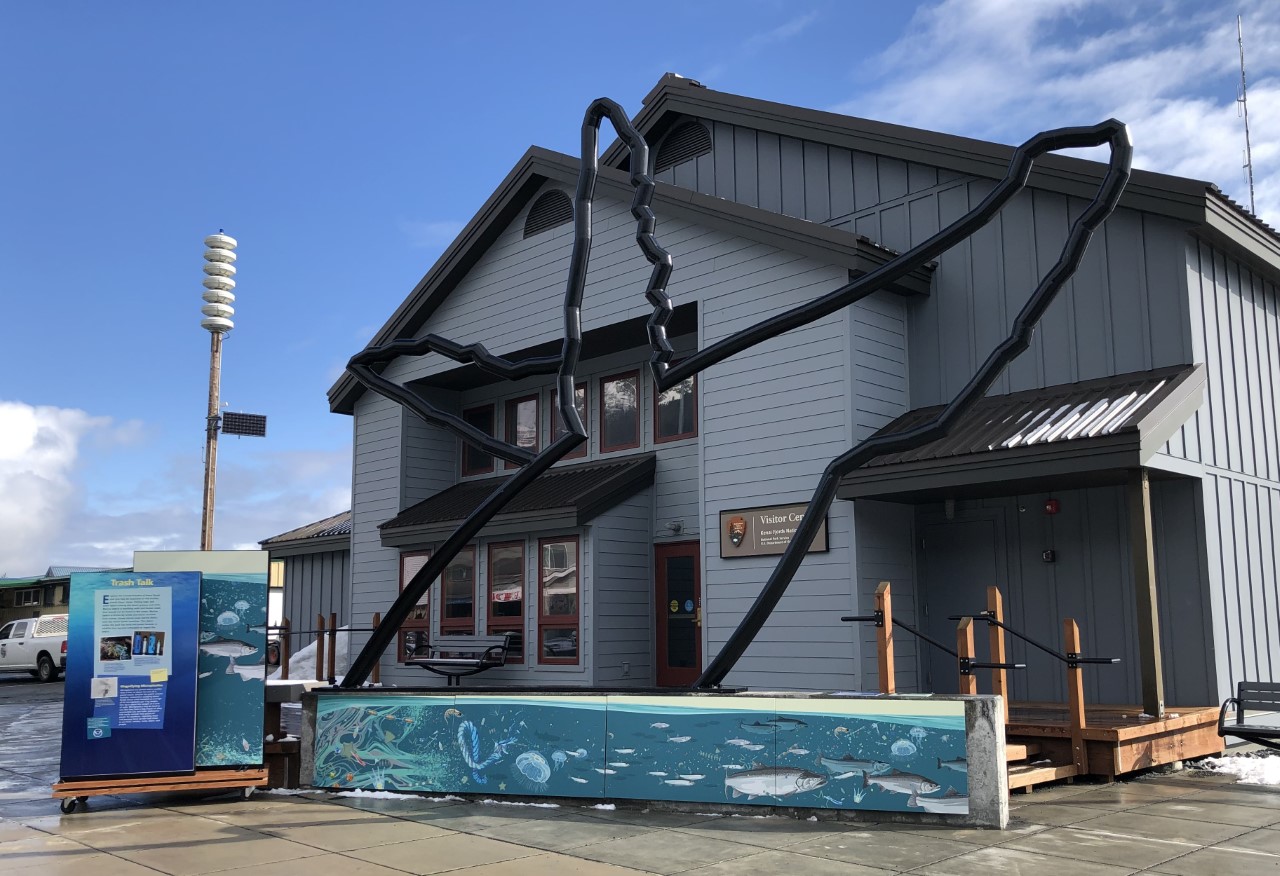 Marine Debris Display at Kenai Fjords a life-size metal sculpture of a whale appears to breach from a low wall with underwater mural painted on it. Next to this is a display on wheels with interpretive panels.