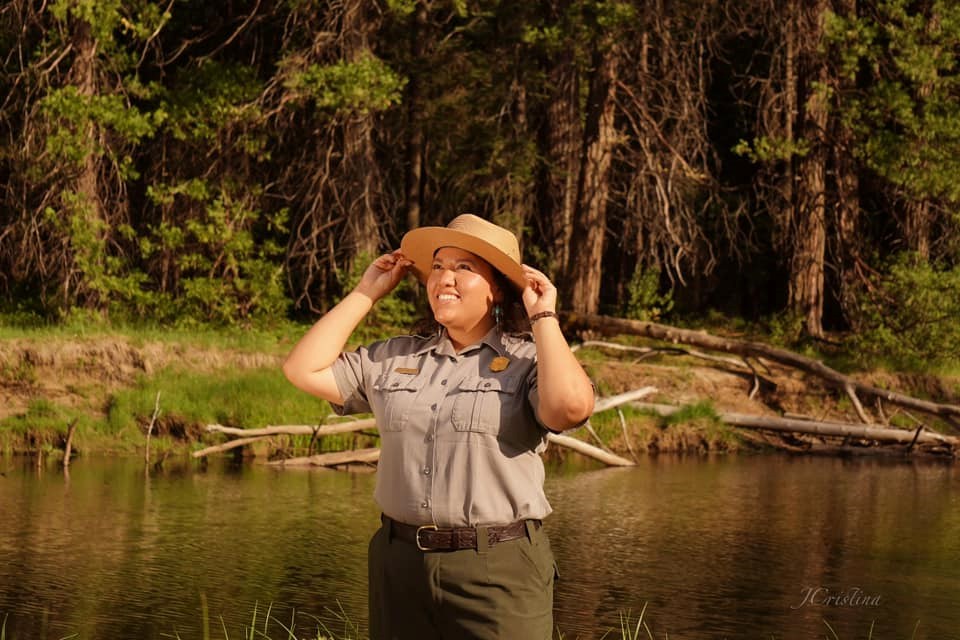 Sandy Hernandez smiles and holds her flathat on her head with both hands. There is a calm creek and trees in the background.