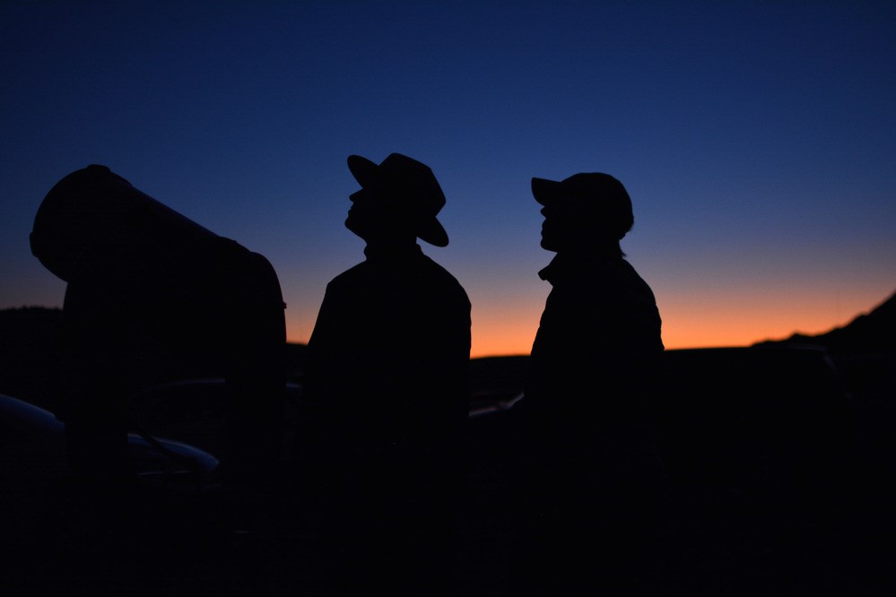 Two silhouetted people, one with an NPS uniform flathat, look up into dark twilight sky beside a telescope