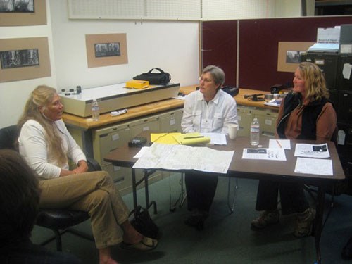 Three people sit at a table with maps, notebooks, and a recorder during an oral history recording