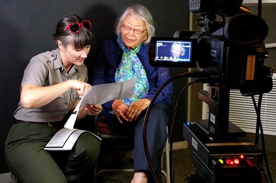 550-MANZ Oral History 201706_GenieObana Genie Obana is seated in front of a video camera, looking at papers with a kneeling woman in NPS uniform