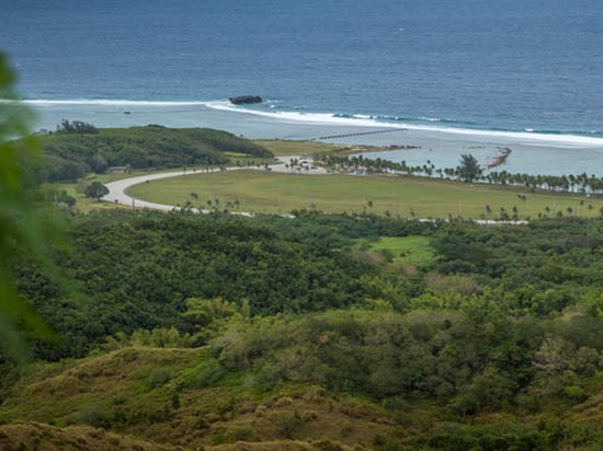 Asan Bay Overlook View from elevation over a lush landscape towards a wide area of turf and palms along a bright blue bay.