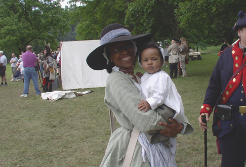 Ajena Rogers, a smiling African American woman holding a child, in a long dress and wide-brimmed hat during living history interpretation.