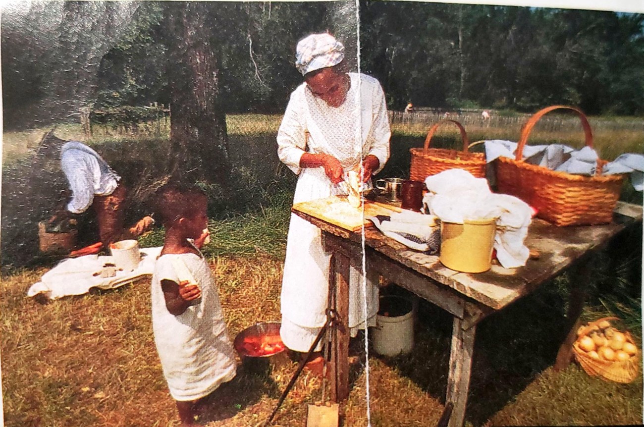 Ajena Rogers, an African American woman and a young boy, her son, stand near a wooden table outside, with woven baskets and other interpretive items.