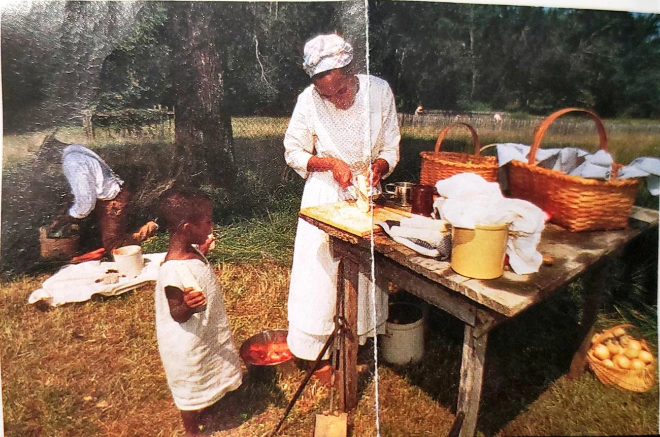 BOWA Ajena Rogers and son Ajena Rogers, an African American woman and a young boy, her son, stand near a wooden table outside, with woven baskets and other interpretive items.