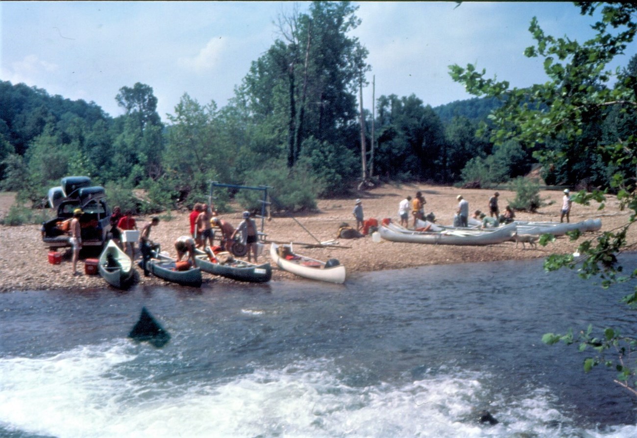 River in foreground and boats, people, and gravel river bank in background.