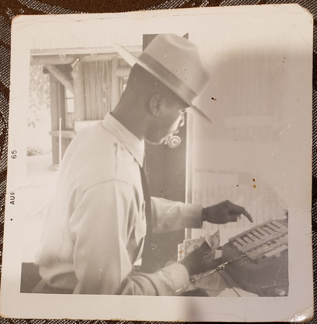 A 1965 photo of a young, African American man in NPS uniform, holding cash and operating a cash register.