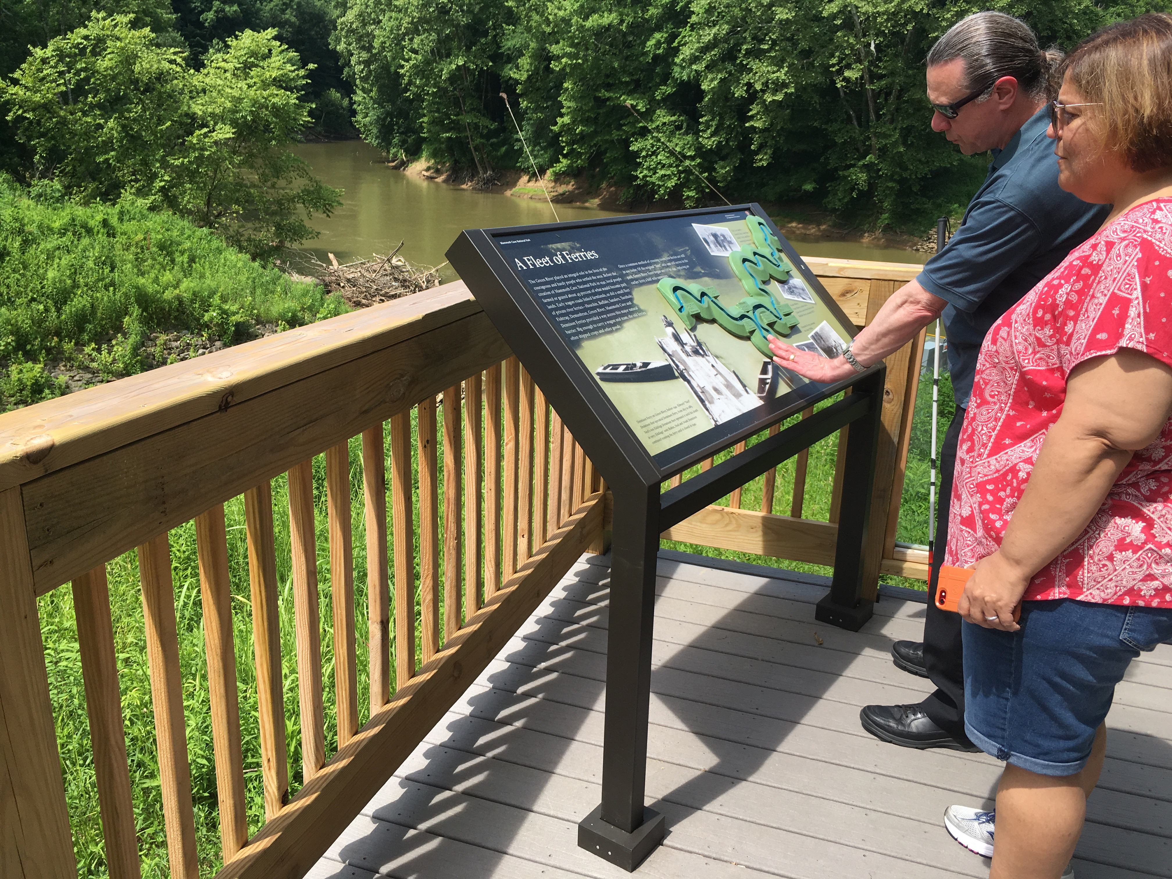 Ray Bloomer touches his fingertips to an interpretive wayside called "A Fleet of Ferries", which stands on a wooden platform overlooking a tree-lined waterway.