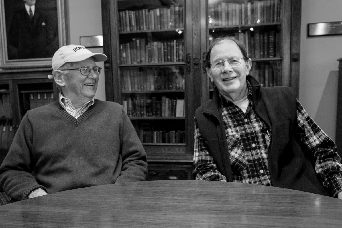 Two men seated at a table, with a bookshelf in the background