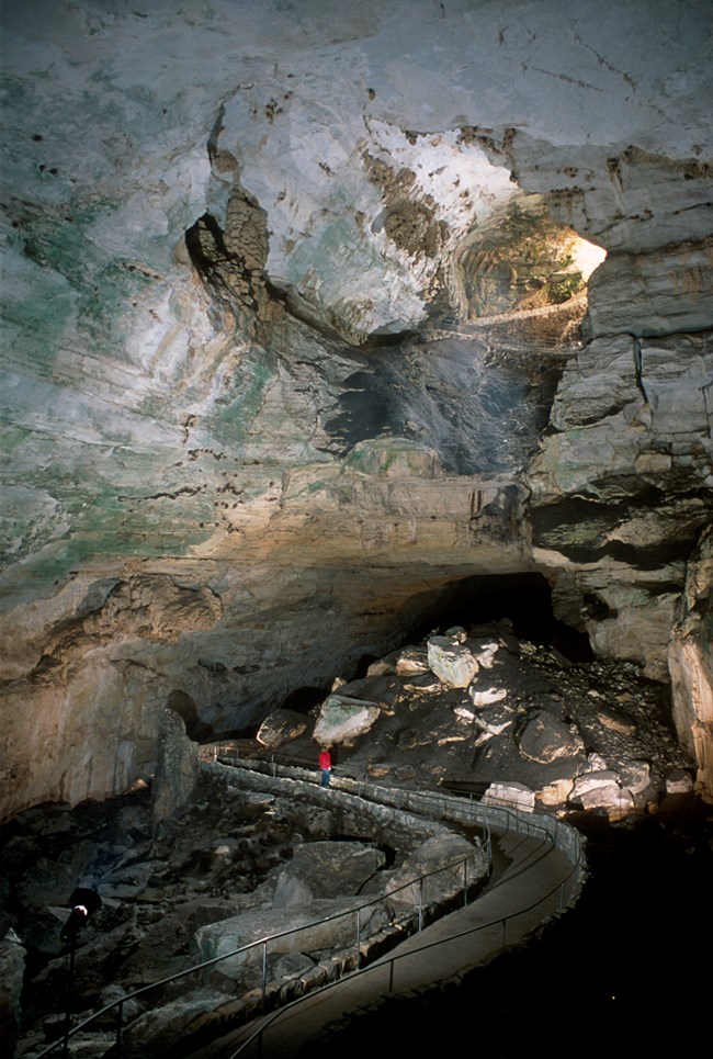 A person, standing on a curving walkway, appears small below the illuminating ceiling of a cavern