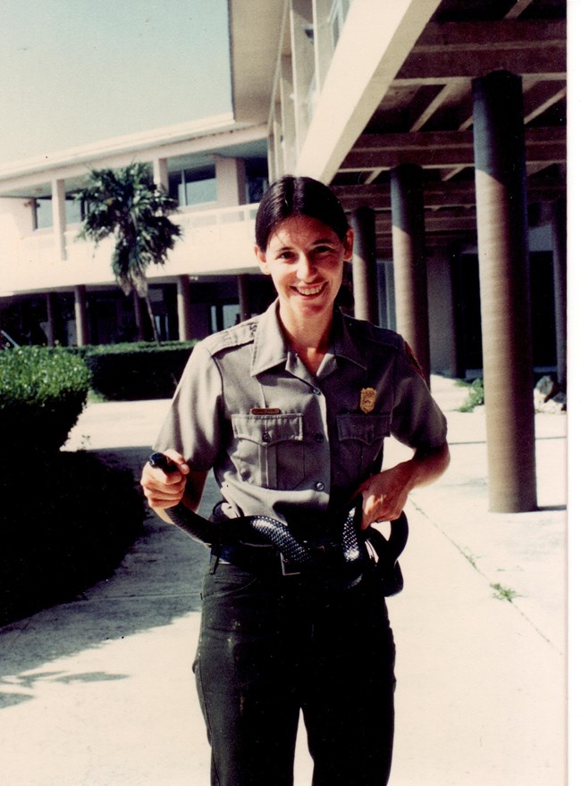 Cherry Payne, in NPS uniform, stands in a couryard beside a building, holding a snake with both hands