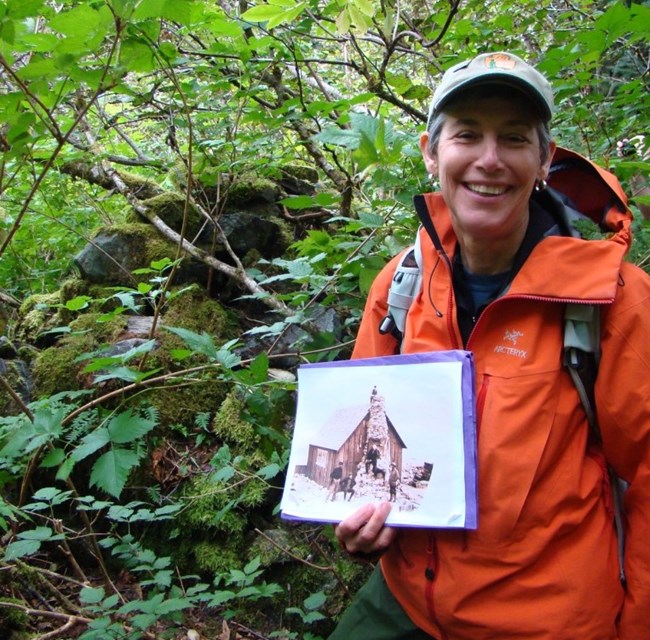 Cherry Payne stands beside a pile of stones covered in vegegation, holding a photographs of a cabin