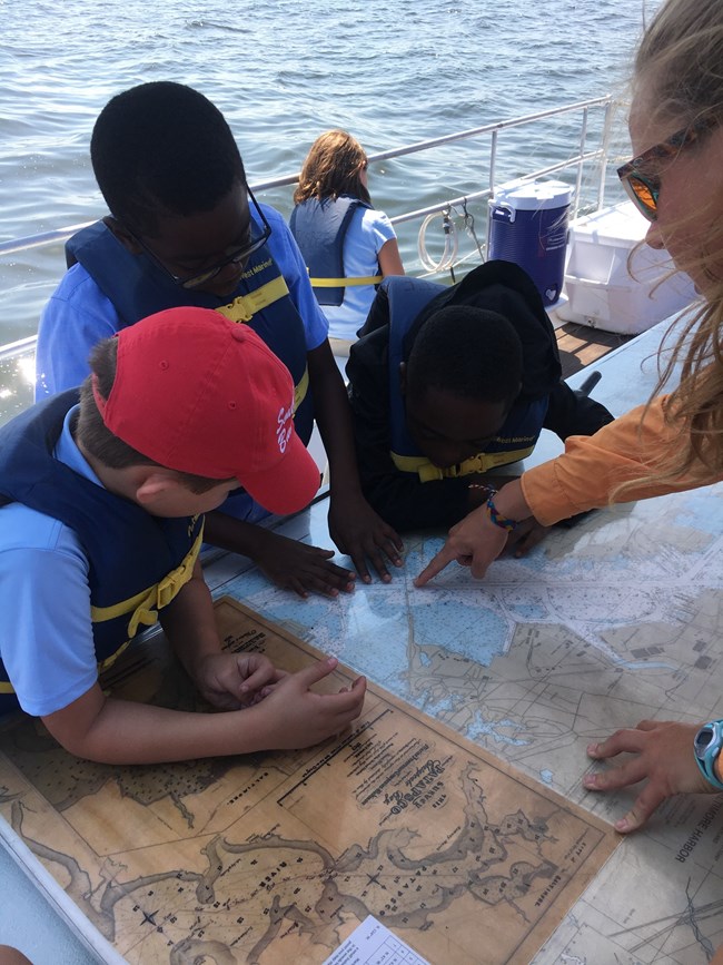 Three children in life-vests lean over to view an area of a map pointed out by a woman, all aboard a boat.