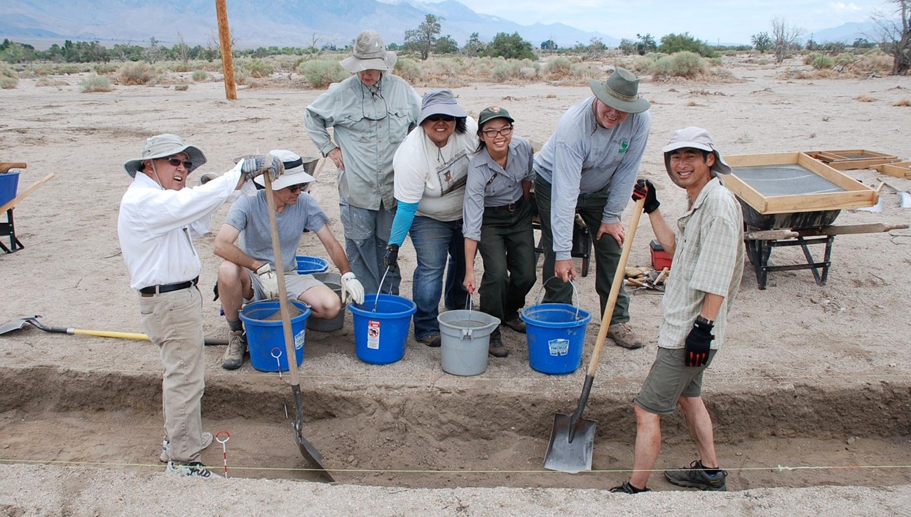 A group of seven people with buckets, shovels, and screenframes stand at the edge of a shallow pit.