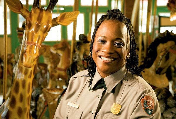 Kym Elder An NPS ranger in uniform, an African American woman, beside on the the animal seats inside a carousel