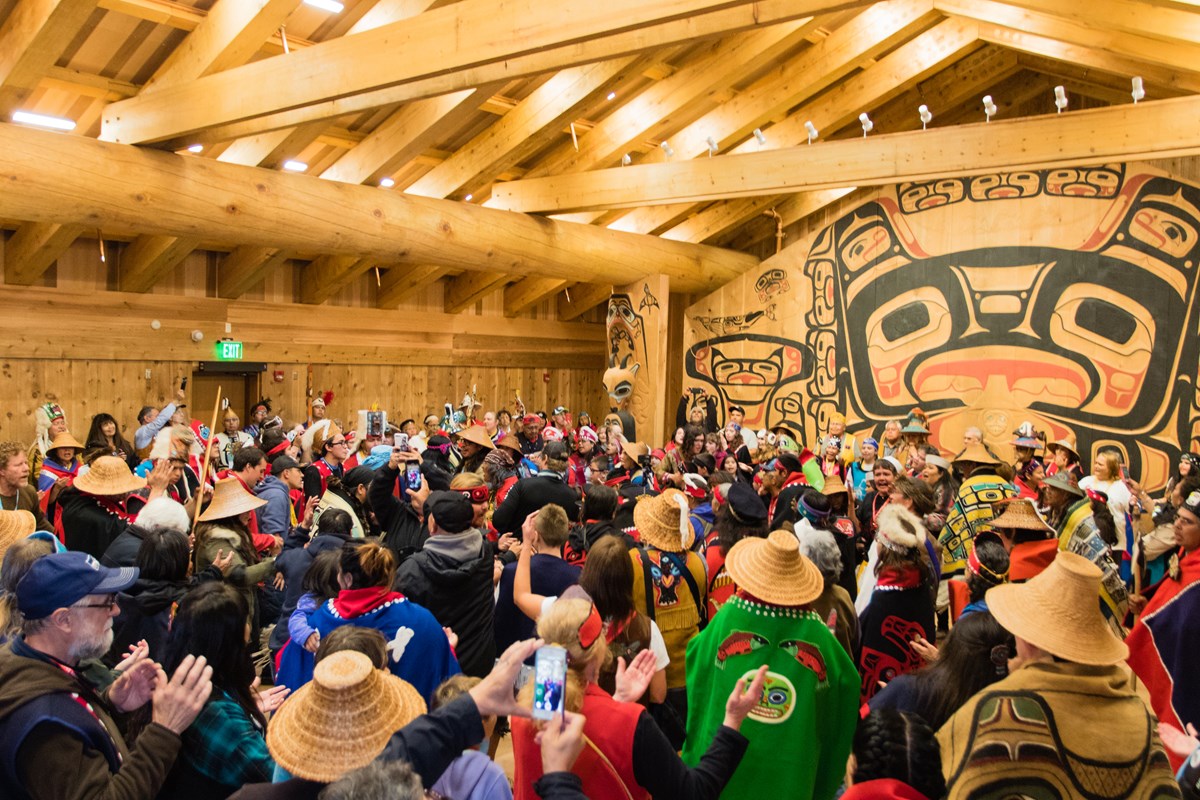 A group of people gather in a large, open building with a high wooden roof and traditional tribal painting on the wall at one end.