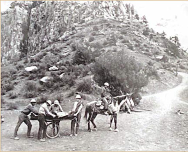 Four people stand around an injured person on a cart pulled by a horse, on a trail at the base of a rocky canyon