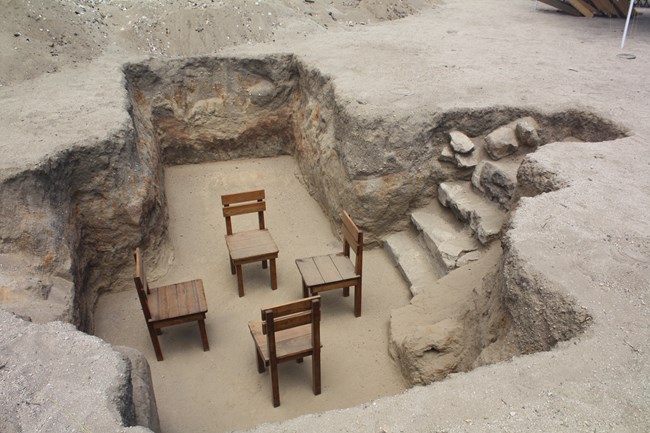 Four wooden chairs face each other in a hole dug into dirt and rock