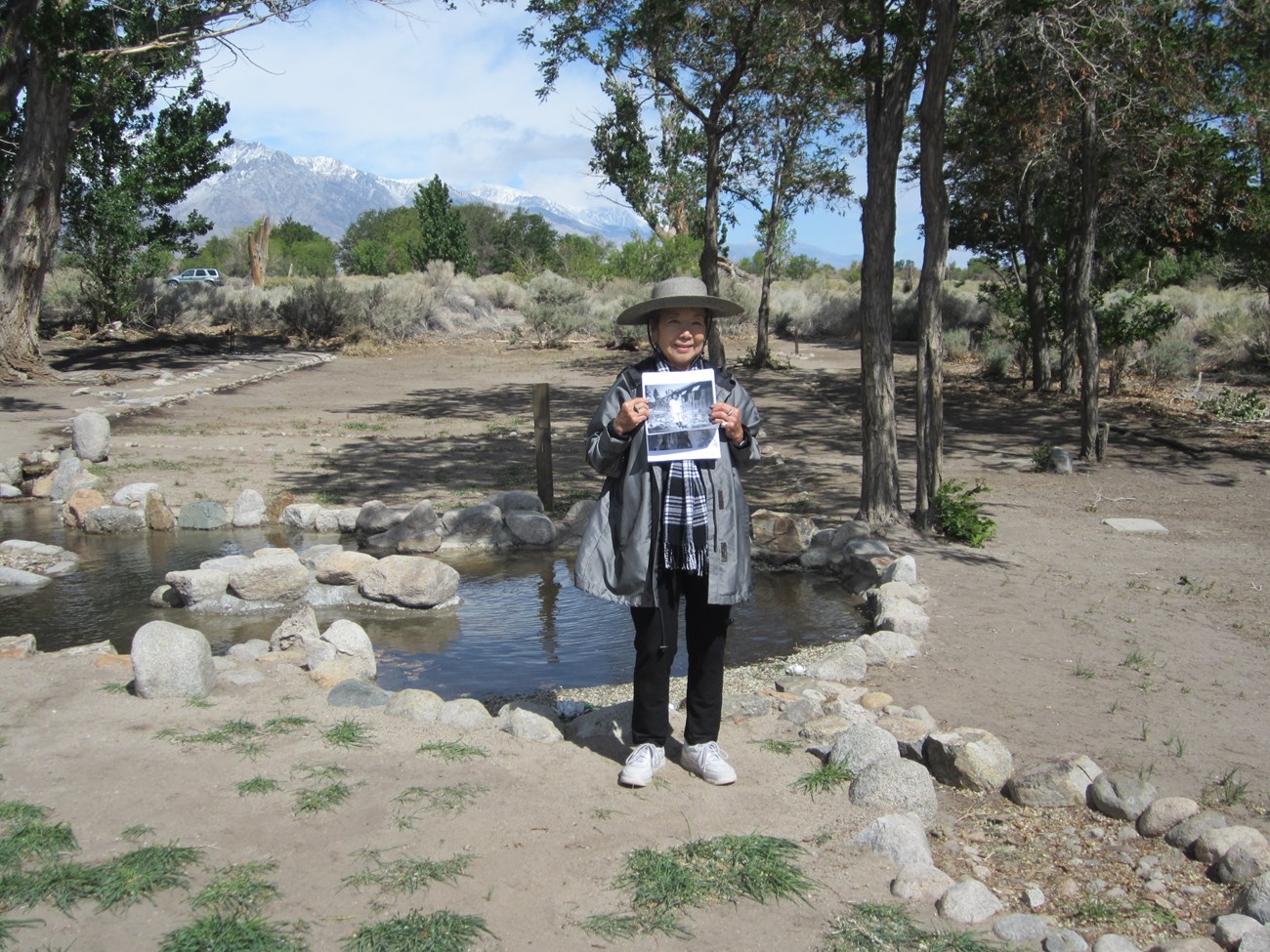 A woman stands in front of a stone-lined pond in an arid landscape, holding a photo of herself as a child in the same location