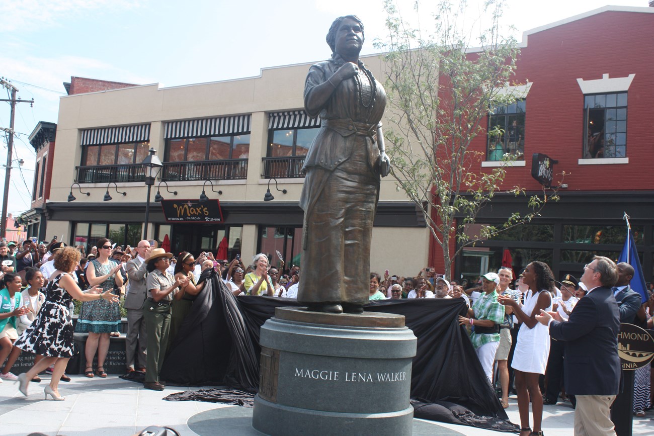 A crowd of people stand around the base of a statue of a woman. The base says "Maggie Lena Walker."