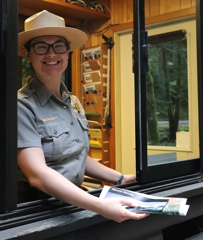 Ranger Emma in NPS uniform outstretches a hand from an entrance station holding a park brochure