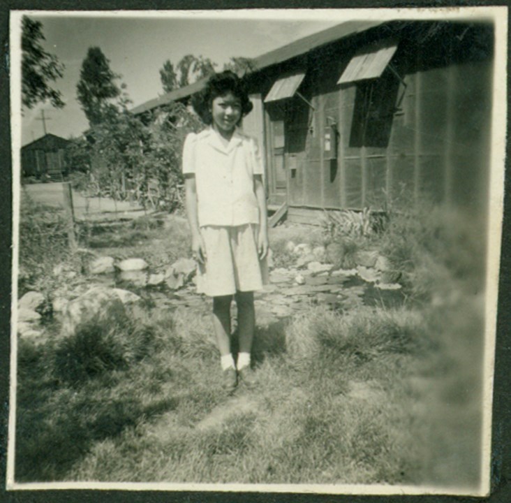 A girl stands in grass beside a small pond, in front of a one-story barracks building. 1944 photo