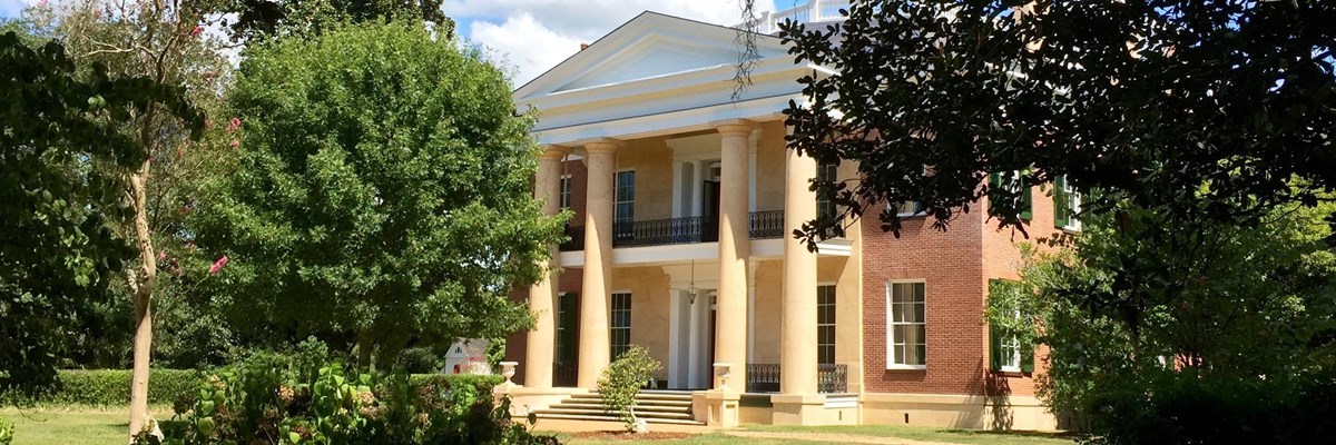 the front of the main house at Melrose Estate has a row of four pillars supporting a porch, framed by leafy vegetation