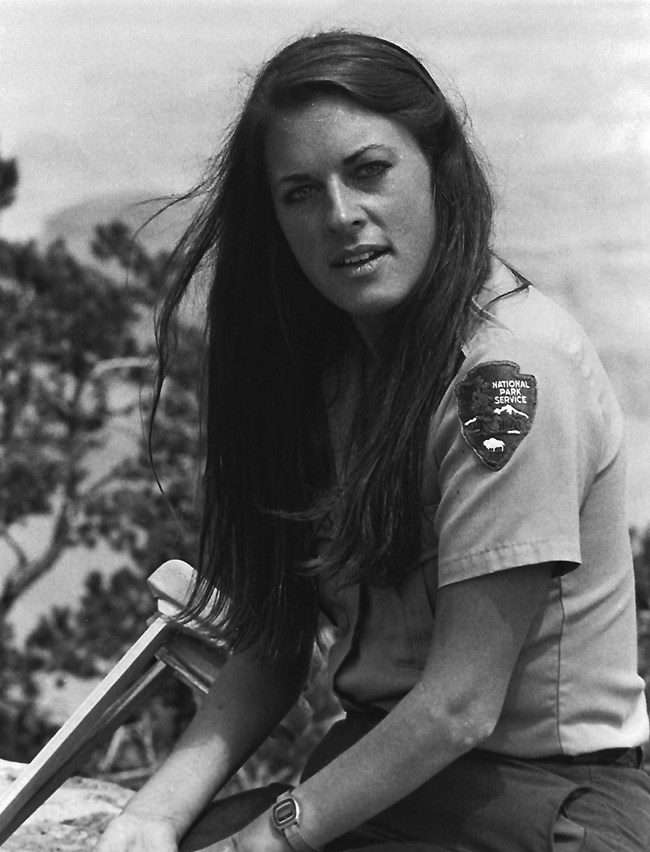 Eileen Szychowski in NPS uniform, seated on a low stone wall with set of crutches beside her.