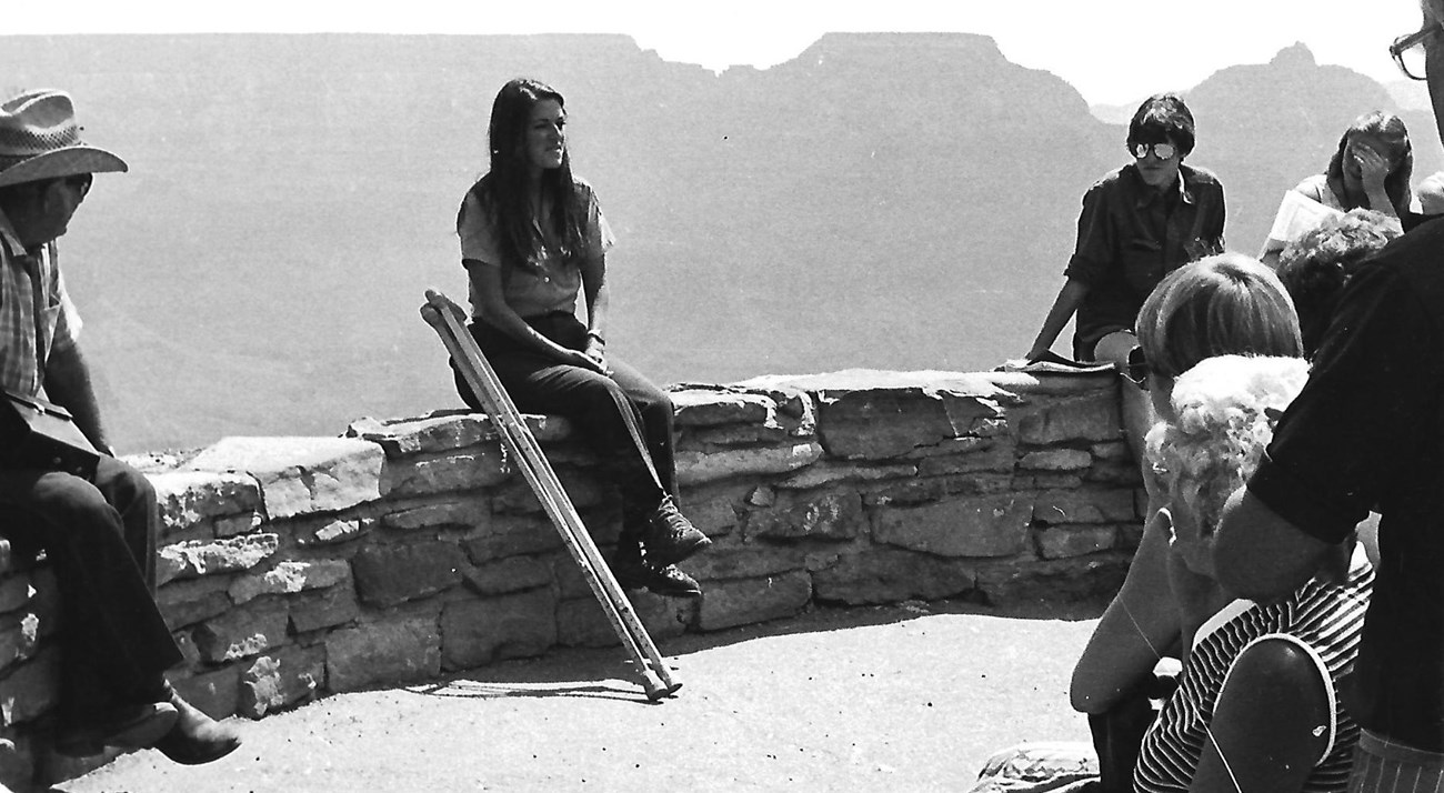 Eileen Szychowski, 1982 Eileen Szychowski, in ranger uniform, seated on a stone wall at the edge of the Grand Canyon with a set of crutches beside her. A group of visitors watches her speak.