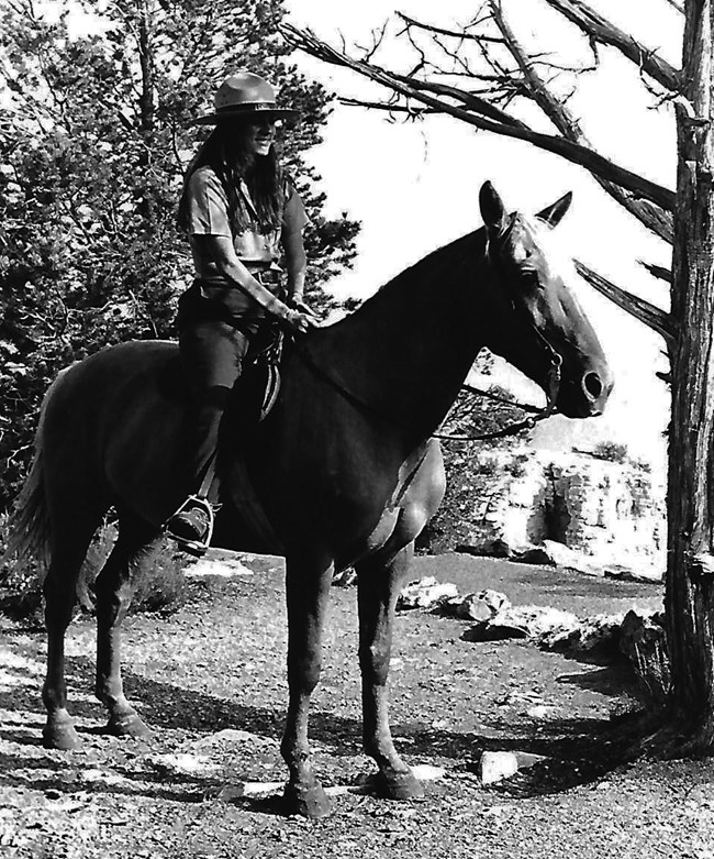 Eileen Szychowski on horseback, 1982 Eileen Szychowski in NPS uniform sits horseback in the Grand Canyon landscape, dry and rocky ground with scattered trees.